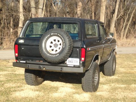 1979 Ford Bronco - The rear-driver side quarter view of a black 1979 Ford Bronco is shown parked on a grassy area with trees in the background. The vehicle fe…