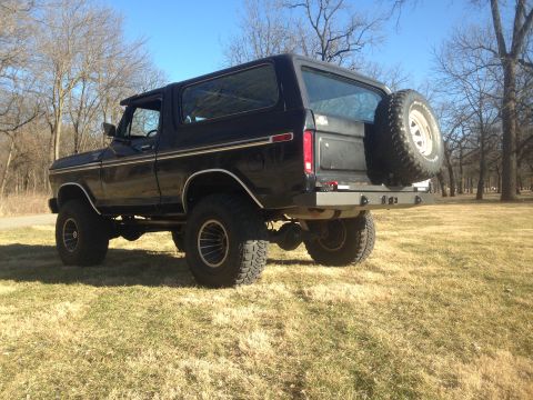 1979 Ford Bronco - The rear passenger side of a black, lifted 1979 Ford Bronco is shown in a grassy field with bare trees in the background. The vehicle featu…