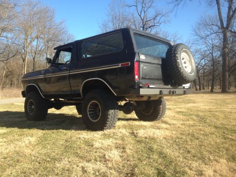 1979 Ford Bronco - The rear passenger side of a lifted black 1979 Ford Bronco is shown parked on a grassy field with bare trees in the background. The vehicle…