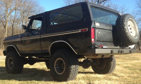 1979 Ford Bronco - The rear driver's side quarter view of a black Ford Bronco is shown parked on a grassy area with bare trees in the background. It features…