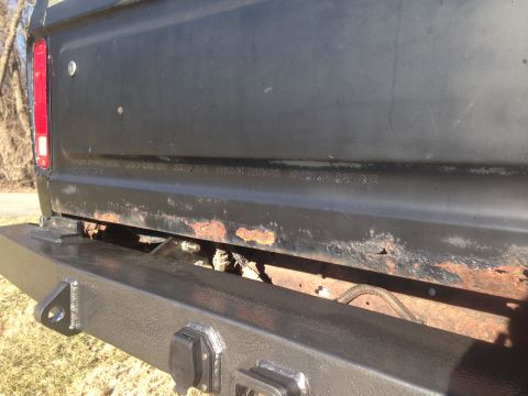 1979 Ford Bronco - Close-up rear view of a black pickup truck tailgate with visible rust and a custom black bumper with trailer connectors. Text is embossed o…