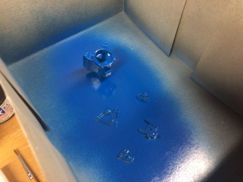 1989 Ford Bronco - Small plastic model parts are being spray-painted blue inside a booth.