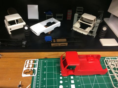 1989 Ford Bronco - Various model car parts, including a red truck cab and bed, are laid out on a workbench next to partially assembled vehicle models.