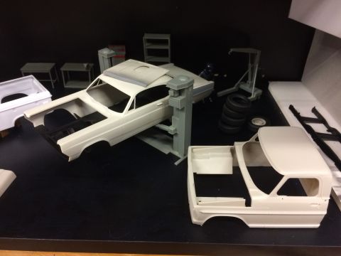 1989 Ford Bronco - Two unpainted model car bodies are displayed on a black surface, along with various model car parts including tires, a car lift, and shelvi…