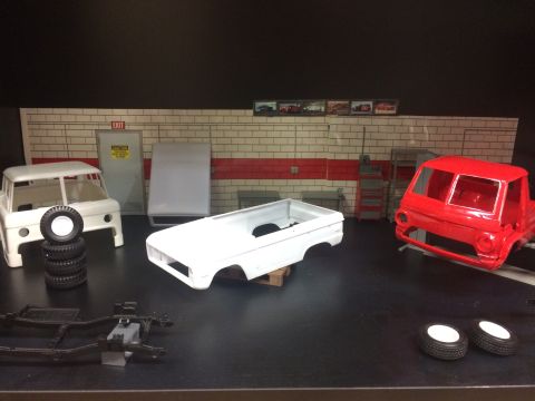 1989 Ford Bronco - Two unfinished model truck bodies, one white and one red, sit on a workbench in a garage diorama. A chassis and wheels are also visible.