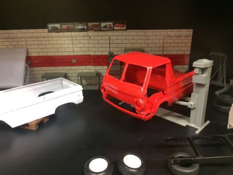 1989 Ford Bronco - A red model truck body is shown on a car lift in a diorama garage setting. Next to it is a white model truck bed on a wooden pallet.