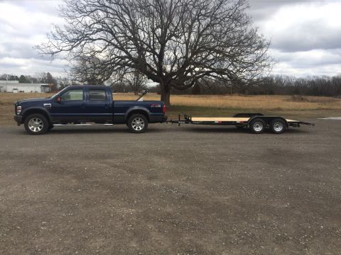 1982 Ford Bronco - A dark blue Ford F-Series pickup truck is parked on a gravel surface, towing a black flatbed trailer. The truck is facing left, with a larg…