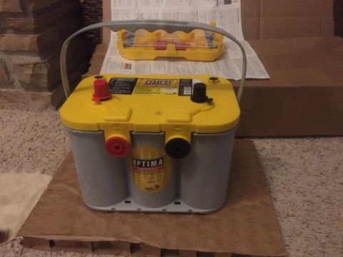 1982 Ford Bronco - A yellow and gray Optima battery sits on a piece of cardboard on a carpeted floor. A yellow plastic battery holder is visible in the backgr…