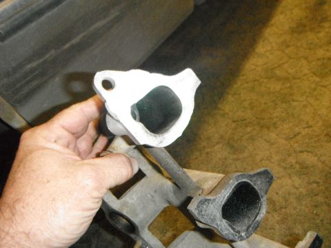 1995 Ford F-150 - A hand holds up a white exhaust manifold gasket against a dark background. Below it, a dark gray exhaust manifold is visible.