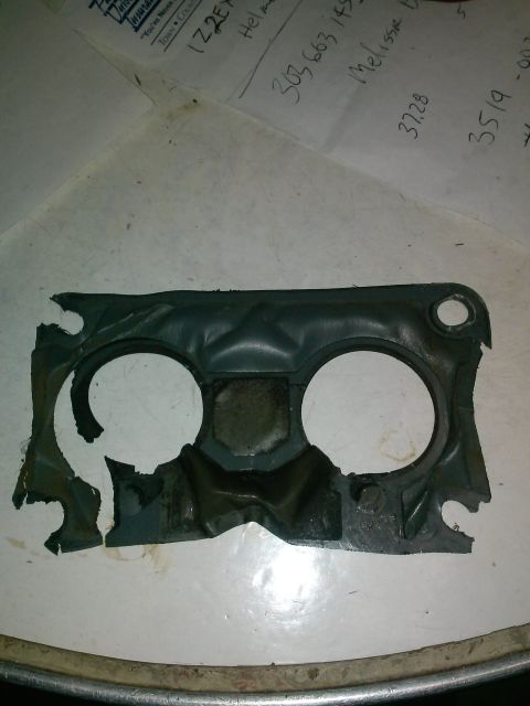 1995 Ford F-150 - A damaged, dark gray gasket with two large circular cutouts and several smaller mounting holes is shown on a white surface. The gasket appe…