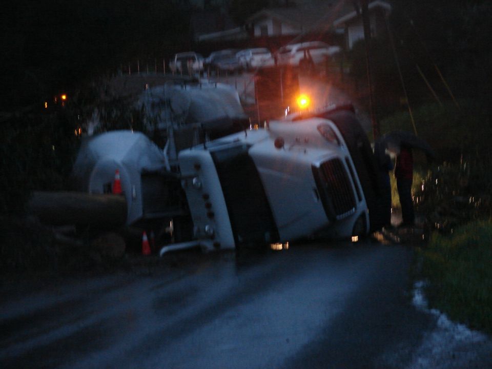 Overturned white tanker truck on a wet road at night with a person holding an umbrella.