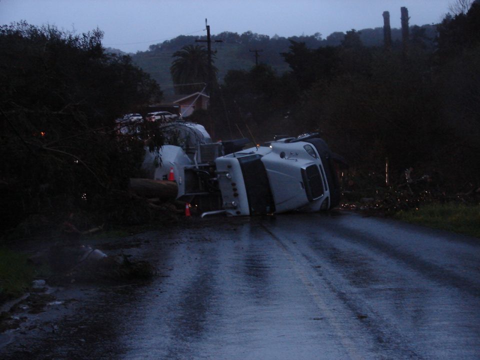 Overturned white semi-truck with logs spilled on a wet road.