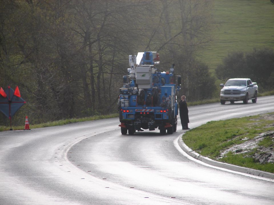 Blue utility truck parked on a wet road with a person standing beside it. Silver pickup truck in background.