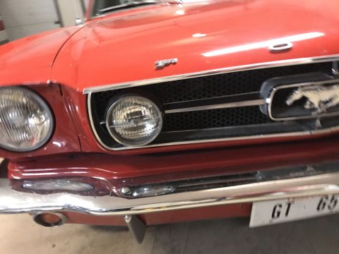 1965 Ford Mustang - The front grille and headlights of a red 1965 Ford Mustang are visible. A silver Mustang emblem is on the grille, and a license plate readi…