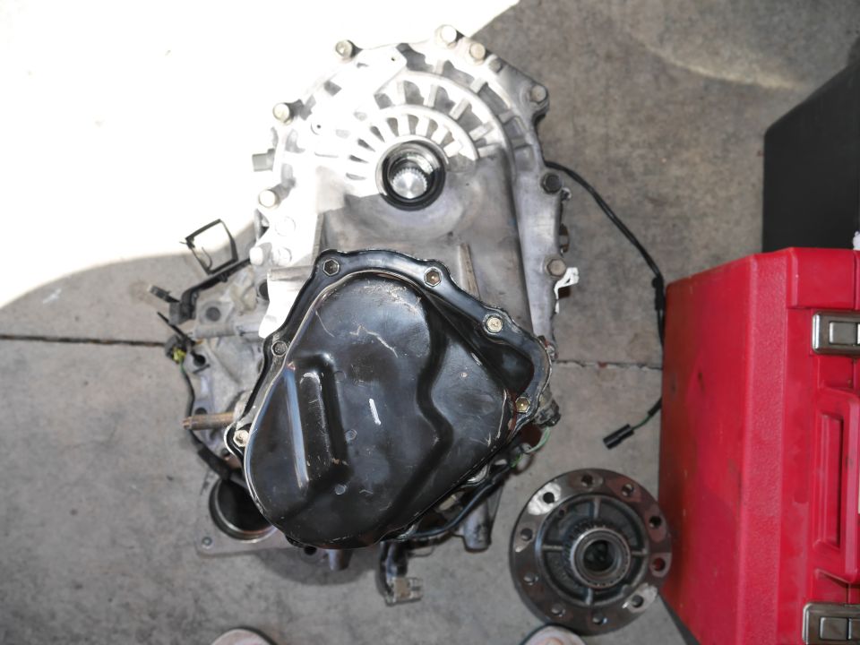 Disassembled automatic transmission with black oil pan next to a red toolbox on concrete.