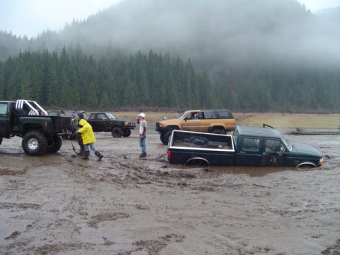1993 Ford Bronco - Off-road vehicles struggle in deep mud near a forested area on a foggy day.