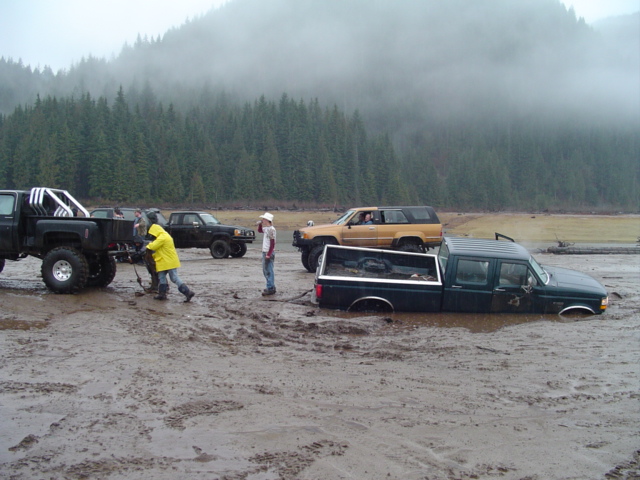 Several trucks are stuck in mud, with people trying to pull one out. Trees and fog are in the background