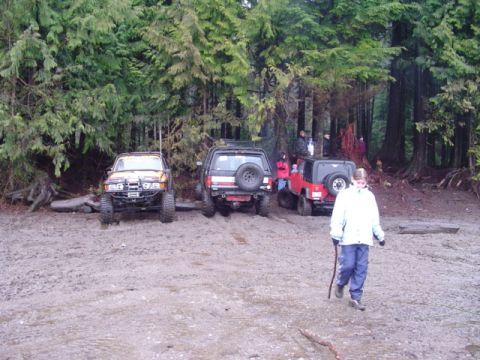 1993 Ford Bronco - Several off-road vehicles are parked in a muddy clearing surrounded by dense trees.