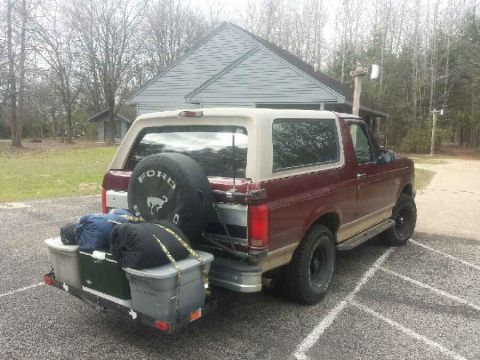 1996 Ford Bronco - A maroon Ford Bronco with a tan top is parked in a gravel lot with a cargo carrier attached to the rear, loaded with storage bins and sleep…