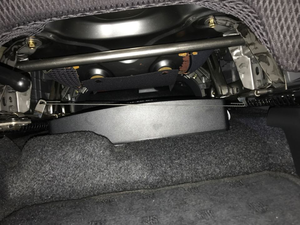 Under car seat, black subwoofer mounted on floor with carpeting and seat frame.
