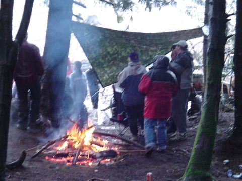 1993 Ford Bronco - A group of people are gathered around a campfire in a wooded area, with a camouflage tarp overhead.
