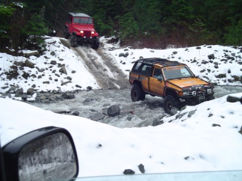 1993 Ford Bronco - Two off-road vehicles navigate a challenging snowy stream crossing in a wooded area.