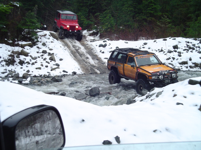 Orange SUV and red Jeep driving through a snowy river, seen from another vehicle's side mirror