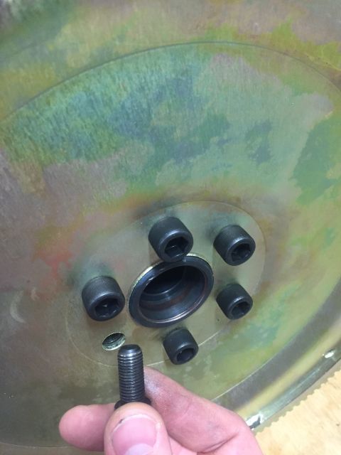 1982 Ford Bronco - A hand holds a bolt in front of a flywheel with six hex bolts installed around a central hub. The flywheel shows signs of heat discoloratio…