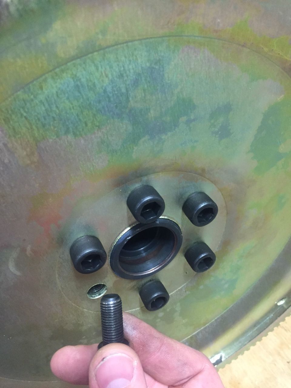 Flywheel with six hex bolts and a loose bolt being held by a hand.