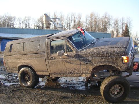 1990 Ford F-150 - This heavily mud-caked Ford F-150, likely a 1990 model, is parked in a muddy lot with a camper shell on the bed.