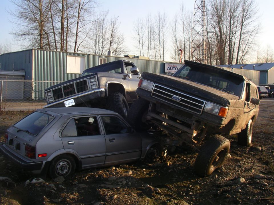 Black lifted Ford truck perched on top of a crushed silver hatchback in a gravel lot outside an auto body shop