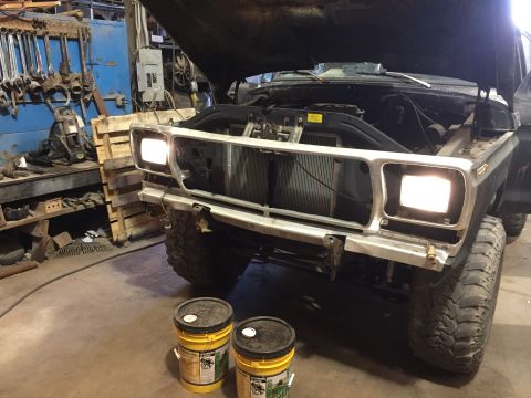 1979 Ford Bronco - The front end of a dark-colored Ford Bronco is shown with its hood open, revealing the engine bay. The headlights are illuminated, and the…