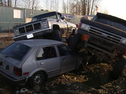 1990 Ford F-150 - A silver hatchback is pinned beneath two large trucks in a junkyard setting.