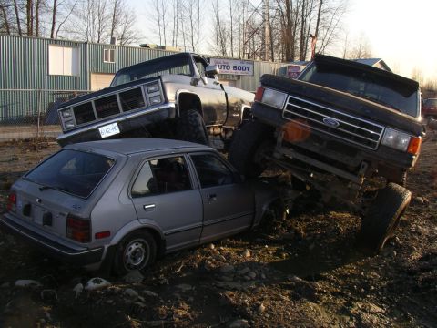 1990 Ford F-150 - A silver hatchback is being crushed by two lifted trucks in a junkyard.