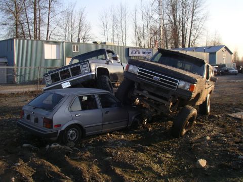 1990 Ford F-150 - A lifted 1990 Ford F-150 is positioned on top of a car, with a GMC truck visible behind.