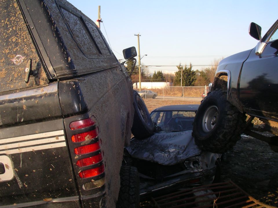 Muddy trucks are piled up, crushing a car between them. One truck is on the left, another on the right