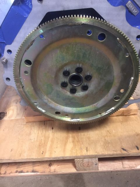 1982 Ford Bronco - A close-up view shows a metal flywheel with a toothed outer edge, bolted to a blue engine block. The flywheel is resting on a wooden pallet.