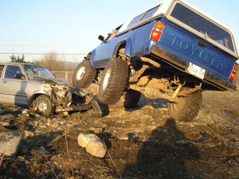1990 Ford F-150 - A lifted Toyota truck is positioned on top of a severely damaged car in a dirt lot.