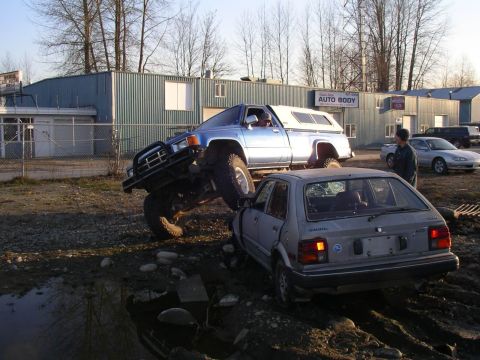 1990 Ford F-150 - A lifted Toyota pickup truck is positioned precariously on top of a silver hatchback outside an auto body shop.
