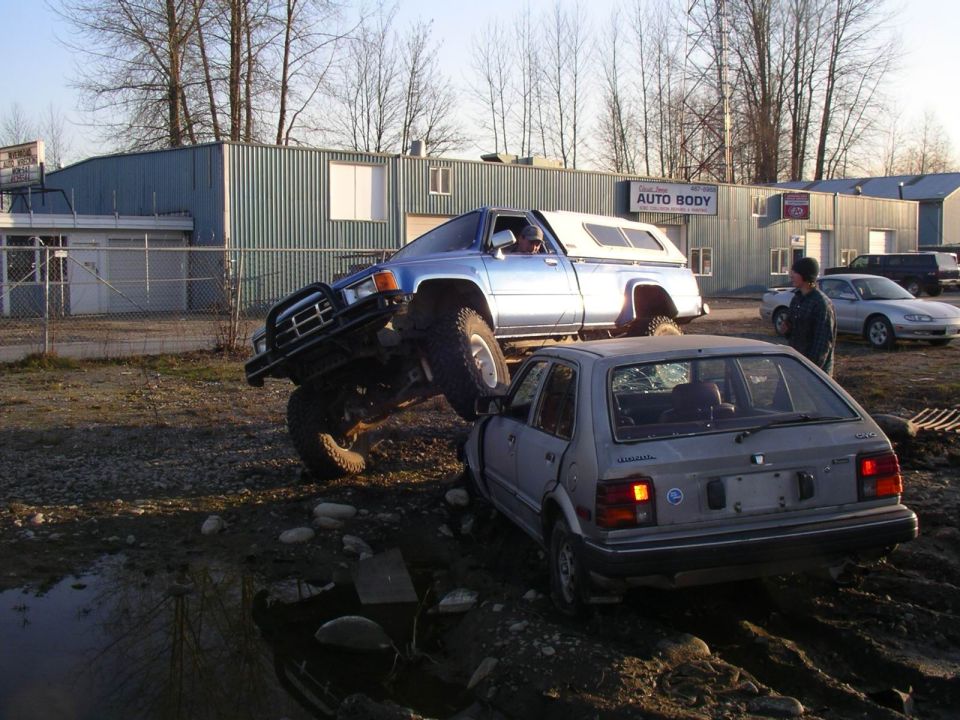 Silver hatchback with broken rear window under a blue lifted Toyota pickup truck. Auto body shop in background