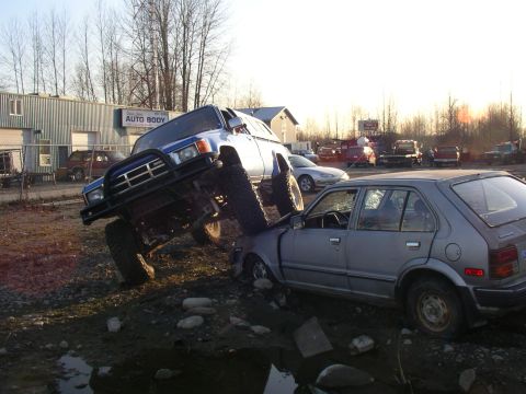 1990 Ford F-150 - A lifted blue Toyota 4Runner is positioned on top of a silver Honda Civic, appearing to crush it, in what looks like a junkyard or auto rep…