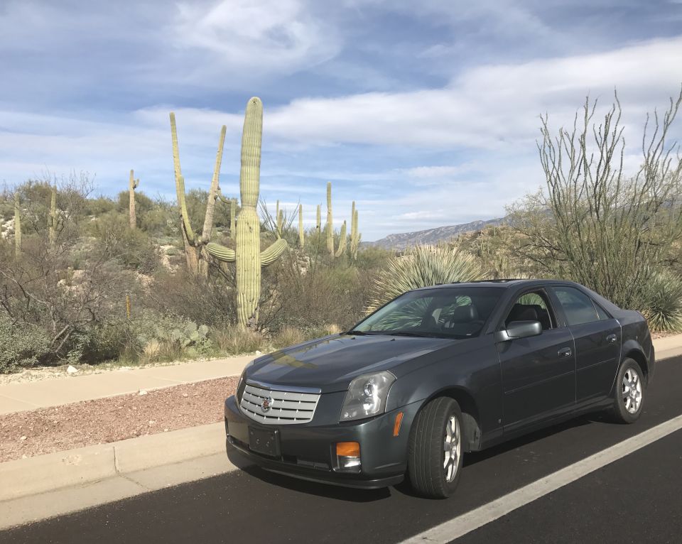 Dark gray Cadillac sedan parked on a road with saguaro cacti and desert landscape.
