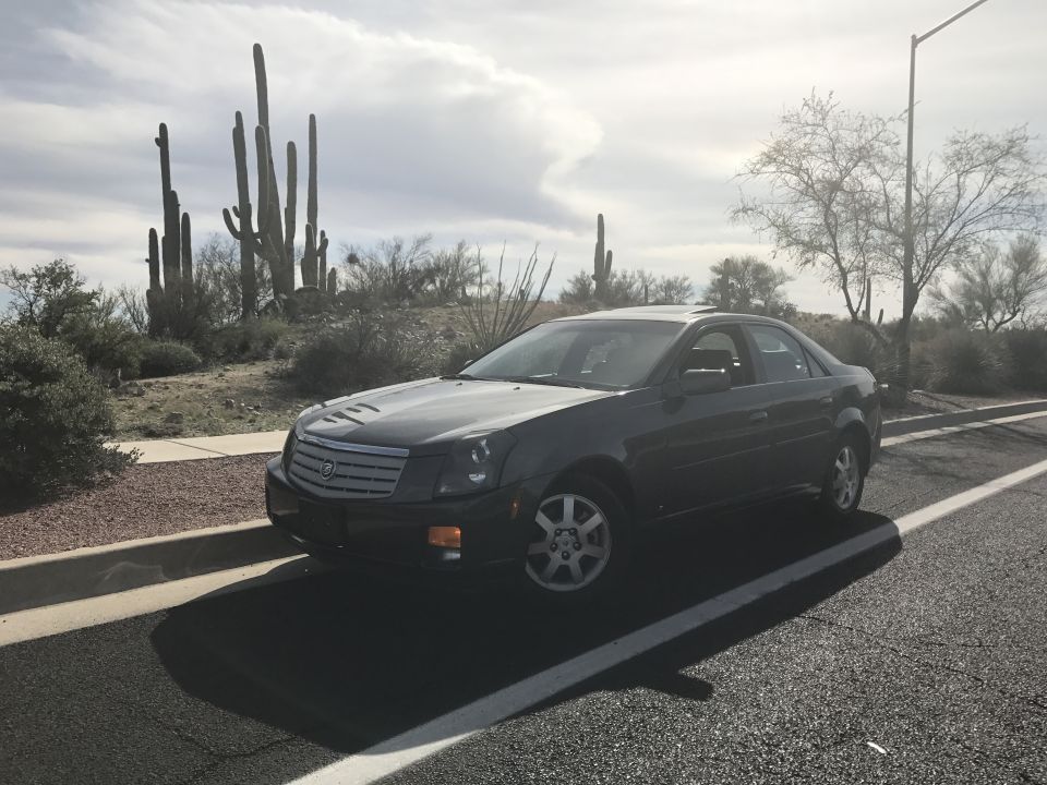 Dark gray Cadillac sedan parked on roadside with desert landscape and saguaro cacti.