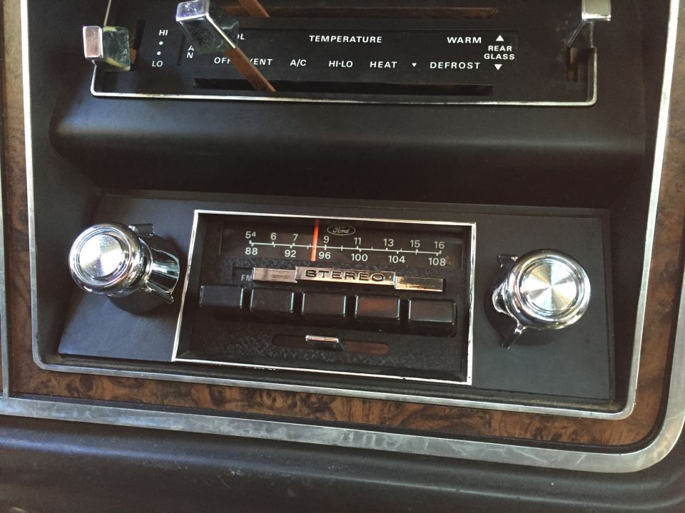 Car dashboard with wood grain trim, a Ford stereo, and climate controls. The radio has a tuning dial and push buttons