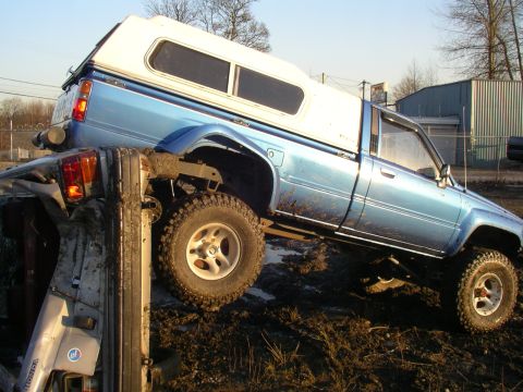 1990 Ford F-150 - A blue pickup truck with a white camper shell is precariously balanced on top of another vehicle in a muddy area.