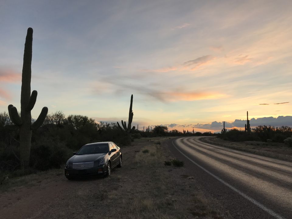 Dark Cadillac sedan parked on dirt shoulder next to a winding road in the desert at sunset with saguaro cacti