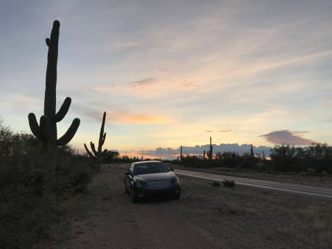 2007 Cadillac CTS - A 2007 Cadillac CTS sits parked on a dirt road in the desert as the sun sets, casting a warm glow on the sky.