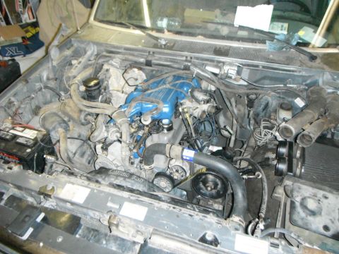 1995 Ford F-150 - The engine bay of a Ford F-150 is shown, featuring a blue intake manifold and various hoses and components. The battery is visible on the l…