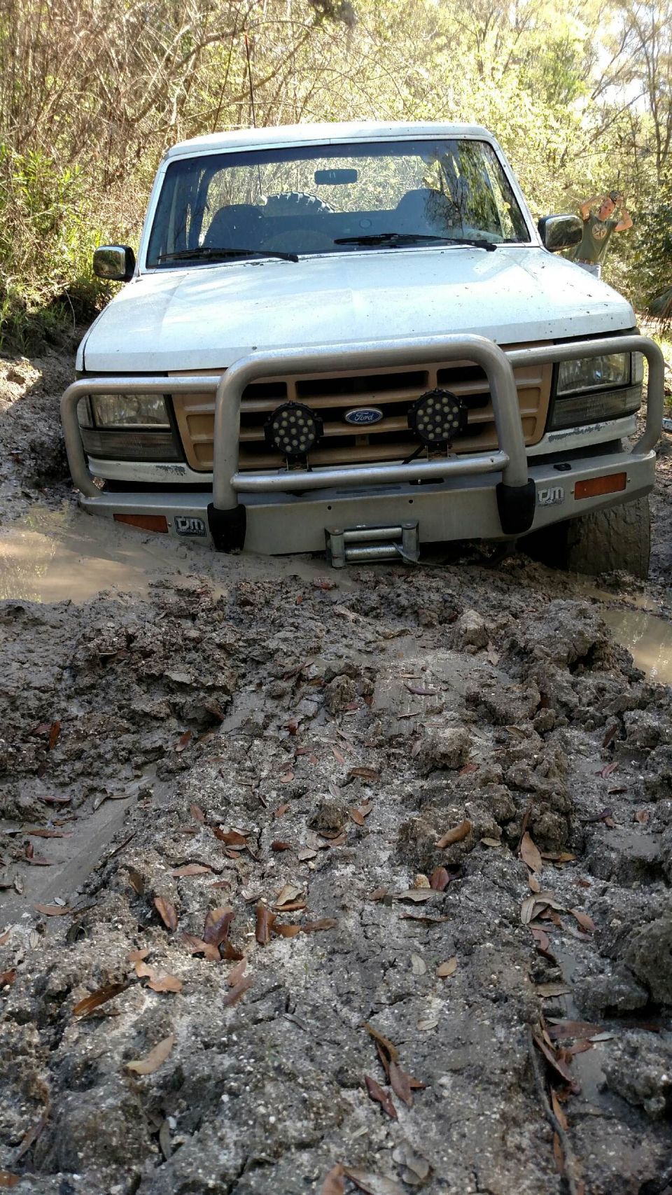 White Ford Bronco stuck in mud on a wooded trail.