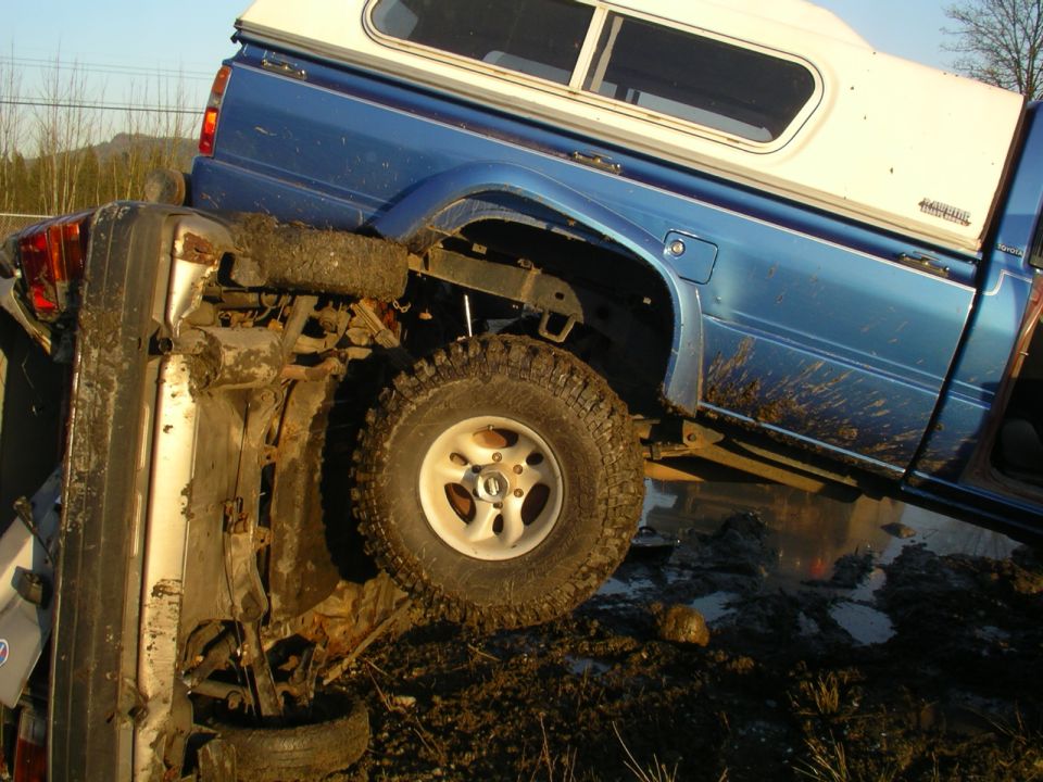 Blue 1990 Ford F-150 pickup truck with a white camper shell, mud-splattered, on top of a silver car